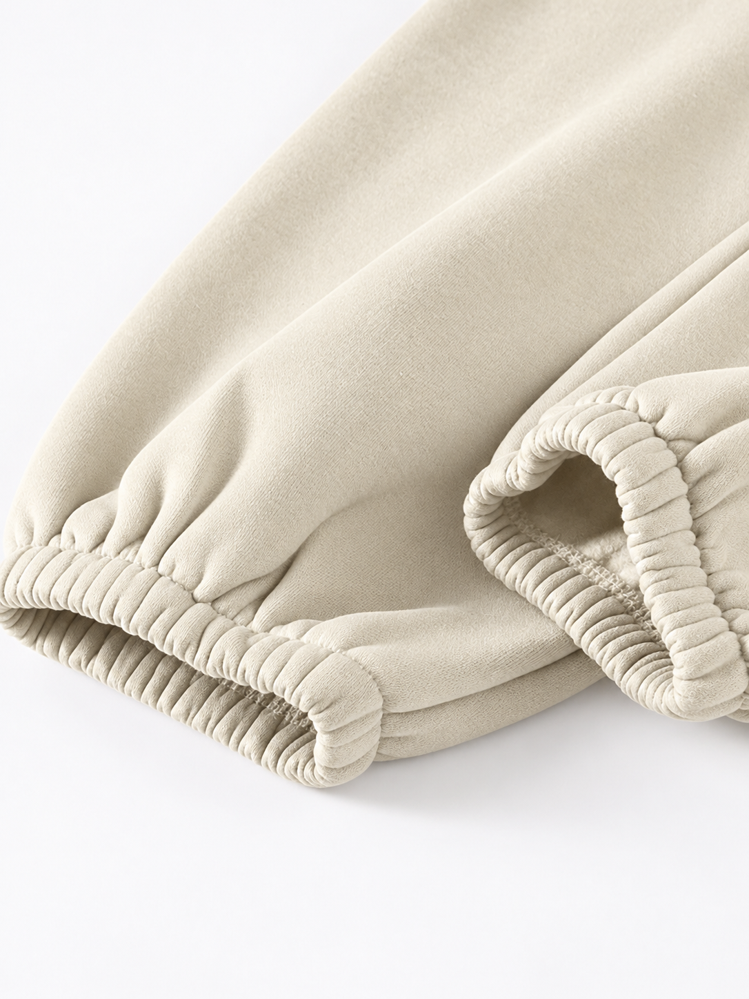 Close-up of a beige fabric garment, likely sweatpants or lounge pants, showing the elasticized ankle cuffs on a clean white background.