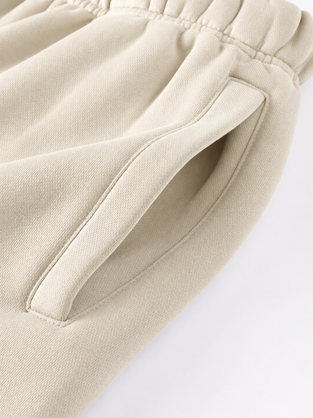 Close-up of a light beige fabric garment showing an elastic waistband and a side pocket with clean stitching on a white background.
