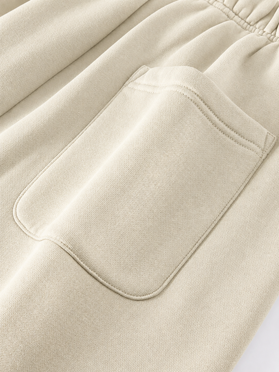 Close-up of a light beige or cream-colored fabric garment, showing a large front patch pocket with neat contrast stitching and an elasticized waistband.